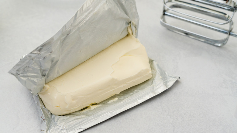 A partially open brick of cream cheese, the foil pulled back, on a grey countertop, the end of a hand-held electric beater behind.
