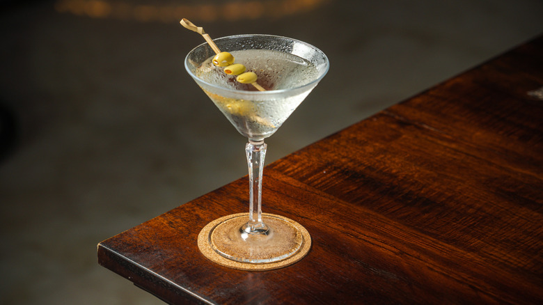 A classic, dirty martini on a coaster, sitting on the corner of a wooden bar.