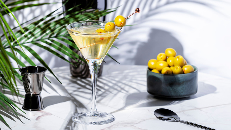 dirty martini, olives, and jigger on a marble counter with a plant frond and shadows in the background