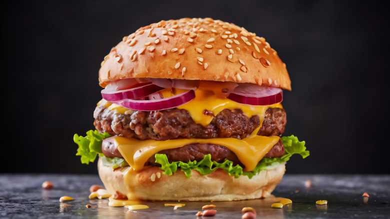 A delicious cheeseburger showcases layers of flavorful beef patties, melted cheese, crisp lettuce, red onions, and sesame seed bun, with a dark background.