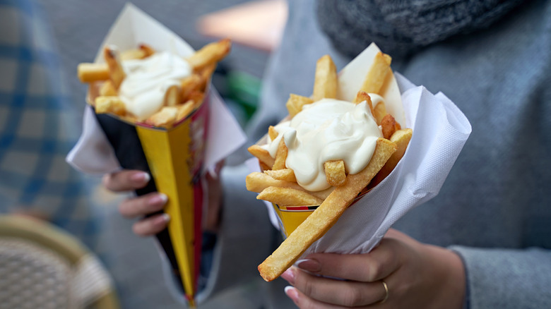 A woman holding two cones of Belgian fries topped with mayonnaise.