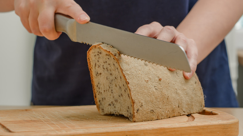 A person's hands are seen cutting a loaf of bread with a large knife.