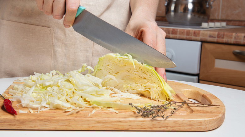 Woman in apron slicing cabbage with sharp knife on domestic kitchen on wooden cutting boars, coleslaw preparation, dinner cooking