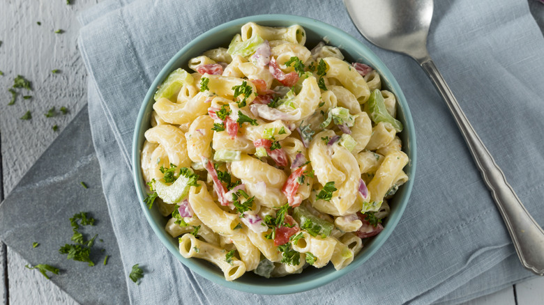 A bowl of macaroni salad next to a metal spoon on a napkin