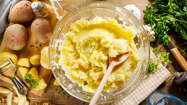 Mashed potatoes in a mixing bowl with potatoes and herbs on the side