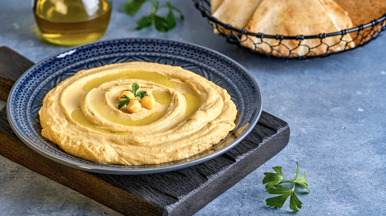 A shallow bowl of hummus on a cutting board with pita and olive oil in the background.