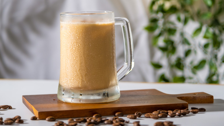 Tall glass mug filled with iced coffee with cream placed on cutting board with coffee beans around it.