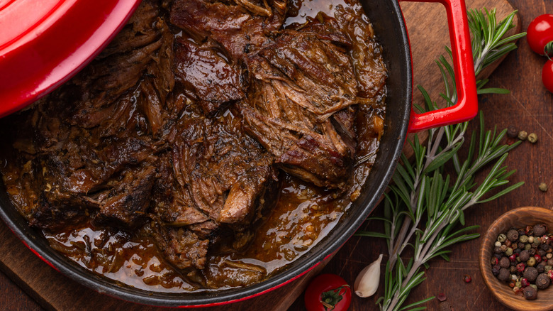 Red dutch oven containing pot roast next to rosemary and peppercorns on wood counter.