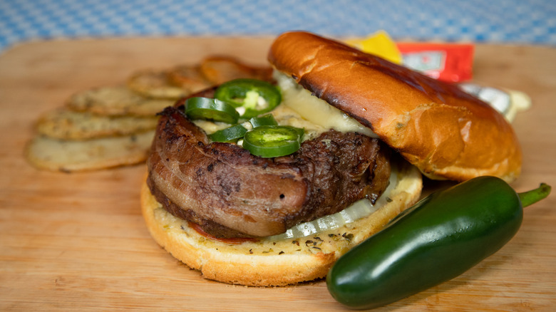 Bacon-wrapped cheeseburger topped with jalapeño peppers, next to a whole pepper on a wooden board