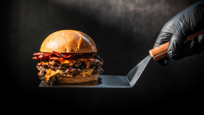 Hand holding spatula with a juicy bacon cheeseburger on it against a black background.