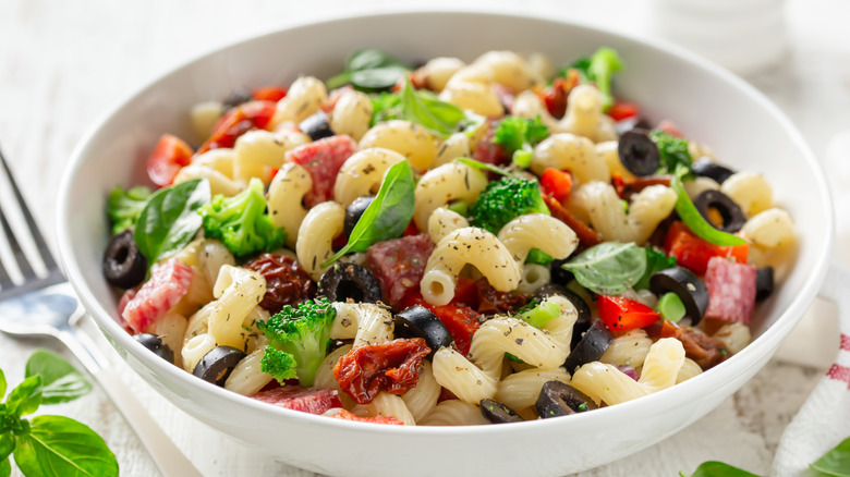 A bowl of pasta salad with basil, salami, olives, and sundried tomatoes