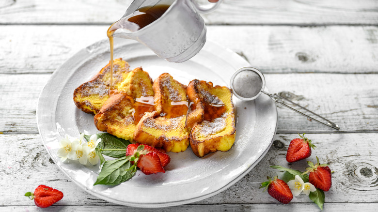 Syrup being poured over French toast with strawberries on a white plate
