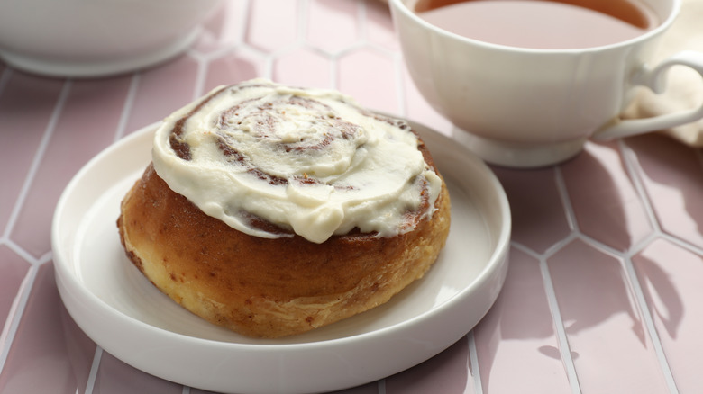 Frosted cinnamon roll on a white plate next to a cup of tea
