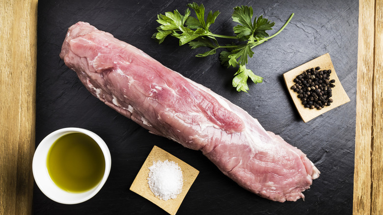 A large pork tenderloin on a slate with salt, pepper, parsley, and oil next to it.