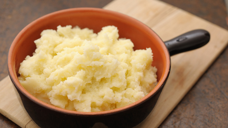 A pot of plain mashed potatoes sits on a cutting board on a table.