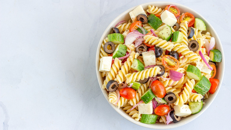 Bowl of pasta salad with olives, cucumbers, tomatoes, and red onion on a white marble counter.