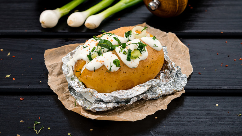 A baked potato with sour cream and chives in aluminum foil on a dark table