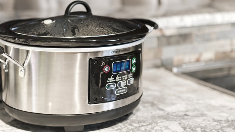 Silver and black slow cooker with a steaming lid on a gray counter