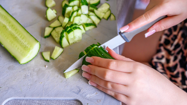 Young hands chopping fresh green cucumber into small pieces on a white cutting board.