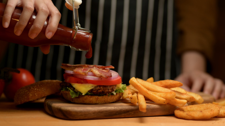 Close up view of female hand with a bottle of ketchup adding on fresh tasty bacon burger in kitchen