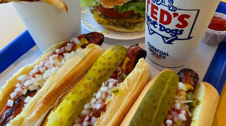 Three hot dogs with onions, pickle spears, and ketchip on tray with cup of fries, burger, and soda
