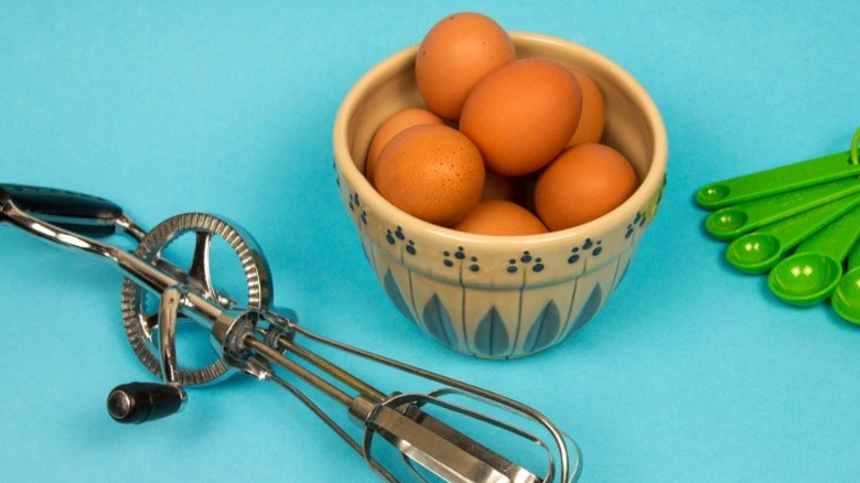 A manual egg beater on a blue background next to a bowl of brown eggs and green measuring cups