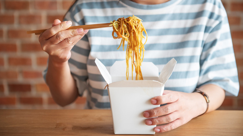 Closeup of hands holding chopsticks, taking noodles out of a white takeout box.