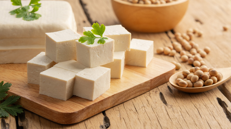 Manicured cubes of tofu are stacked on a wooden cutting board amid herbs and soybeans.