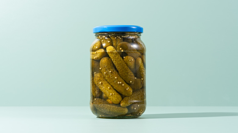 A glass jar of pickled gherkin cucumbers with a blue top on a light green background over a light green countertop,