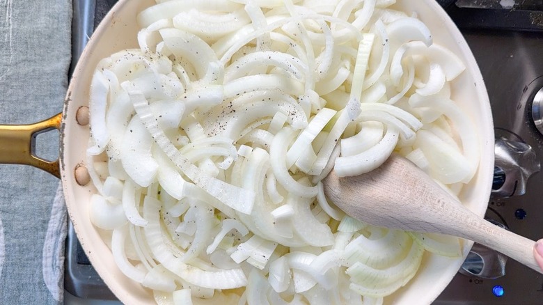Stirring sliced yellow onions with salt and pepper in a large skillet with wooden spoon