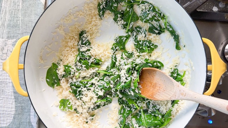 Stirring spinach leaves into white rice grains in large pot with wooden spoon