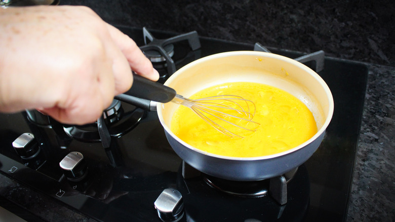 A person's hand is seen whisking scrambled eggs on a stovetop.