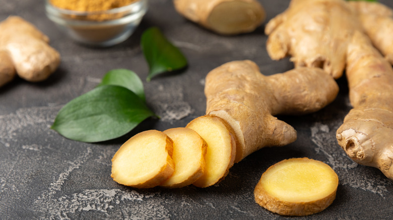 Sliced fresh ginger root on a black countertop next to leaves and a bowl of dried ginger