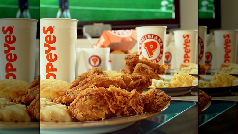 Plate of Popeye's fried chicken with biscuit and mashed potatoes, cups in the background, and a TV playing sports
