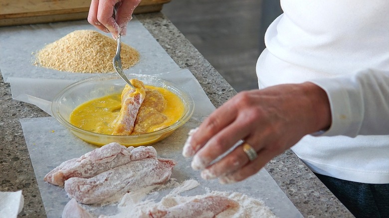 Hands dredging raw chicken in flour, egg, and breadcrumbs over a long sheet of parchment paper