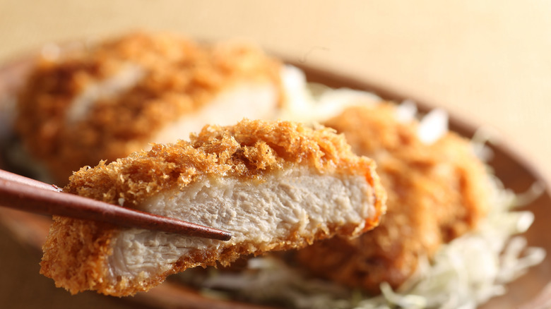 A piece of chicken katsu held in chopsticks above a bowl.