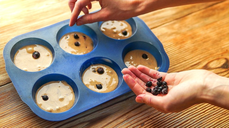 Hands adding frozen blueberries to raw batter in a six-count muffin tin