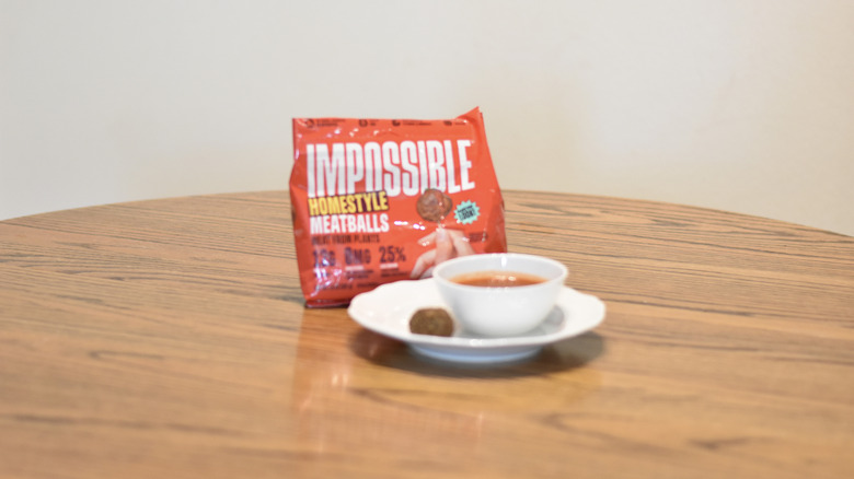A bag of Impossible homestyle meatballs next to a meatball and marinara on a white plate