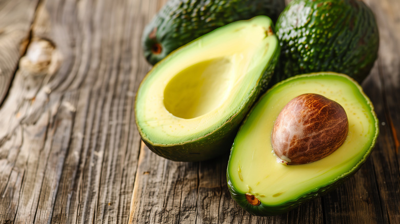 Two avocado halves on a wooden table with two whole avocados in the background
