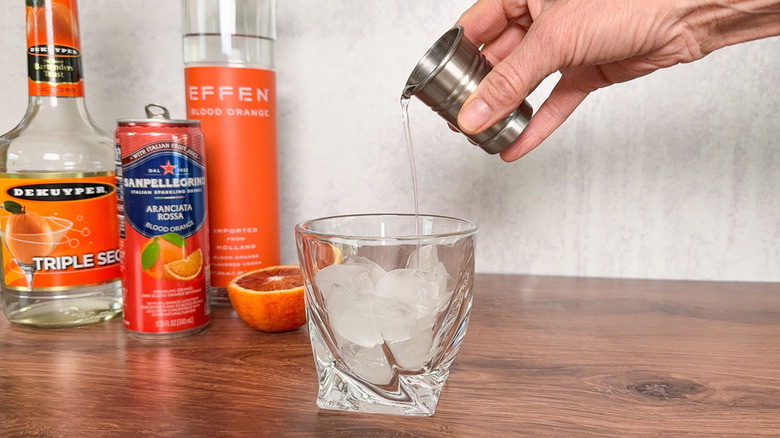 Pouring shot of vodka over ice in rocks gladd with blood orange vodka and soda and triple sec bottles