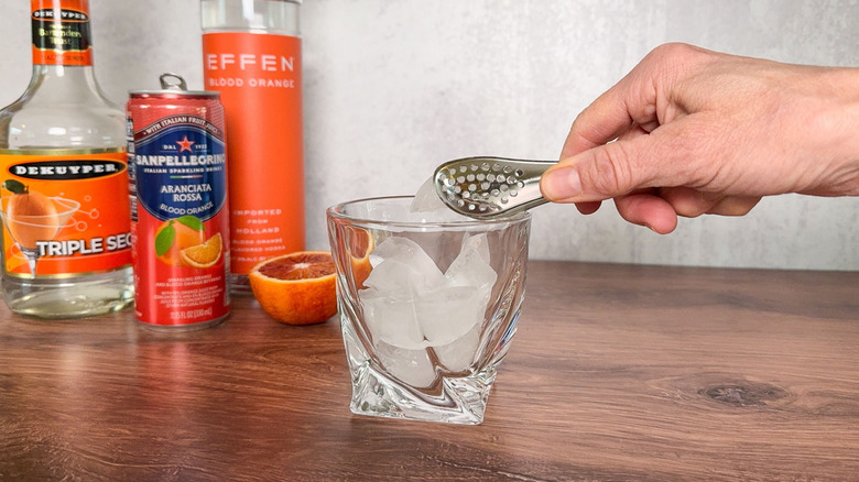 Filling a rocks glass with ice using tongs and blood orange vodka, soda, and triple sec in background