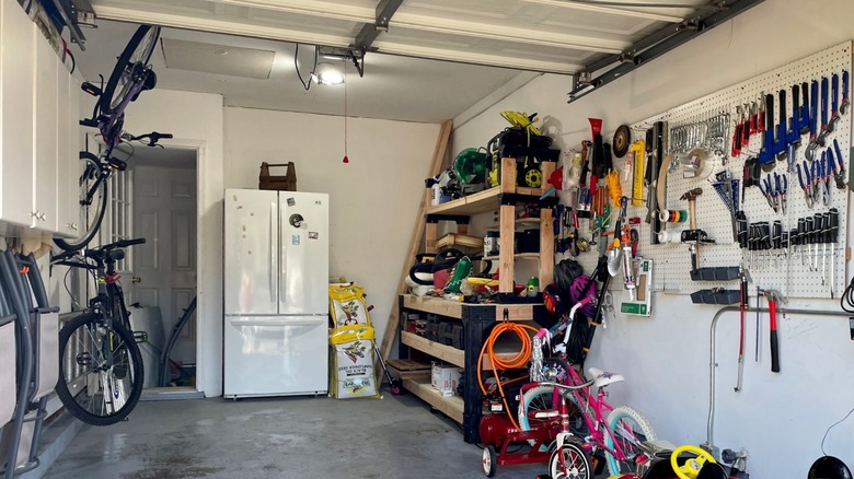 A white refrigerator inside of a typical household garage with tools, bikes, garden supplies, and clutter.