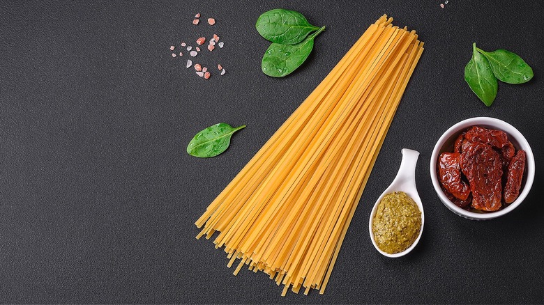 A stack of dried spaghetti noodles, fresh basil leaves, and a bowl of sun-dried tomatoes