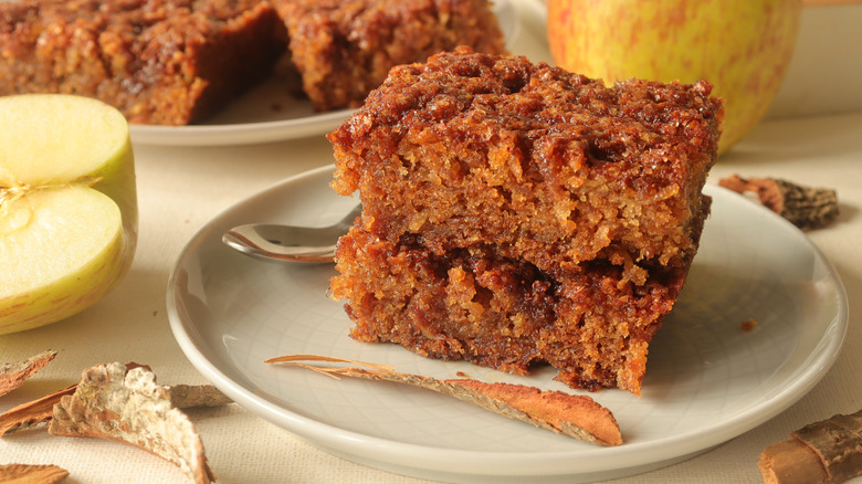 an apple caramel cake sitting on a white plate, apples in the background and a spoon behidn it
