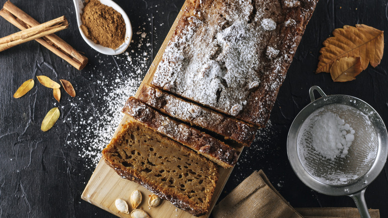 Gingerbread loaf on cutting board with powdered sugar and spices on black table