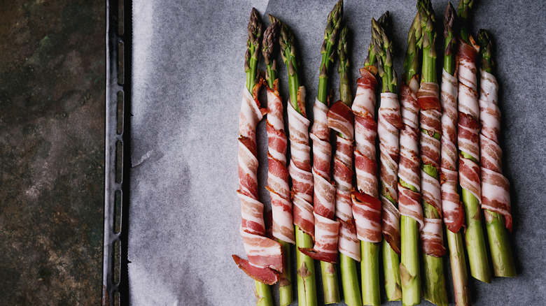 Asparagus wrapped in bacon, arranged on a baking tray lined with parchment paper.