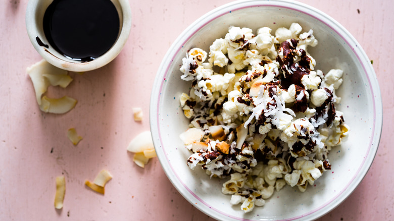 Bowl of popcorn topped with chocolate and coconut next to a cup of melted chocolate