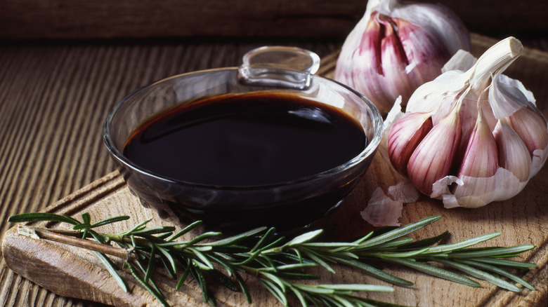 A small glass bowl of balsamic beside whole garlic globes and herbs.