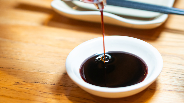 A close-up of soy sauce being poured from a dispenser into a small white dish, with chopsticks in the background