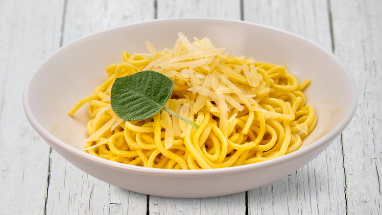 Sage-topped spaghetti pasta with butter and parmesan shreds in a white dish on white wooden table